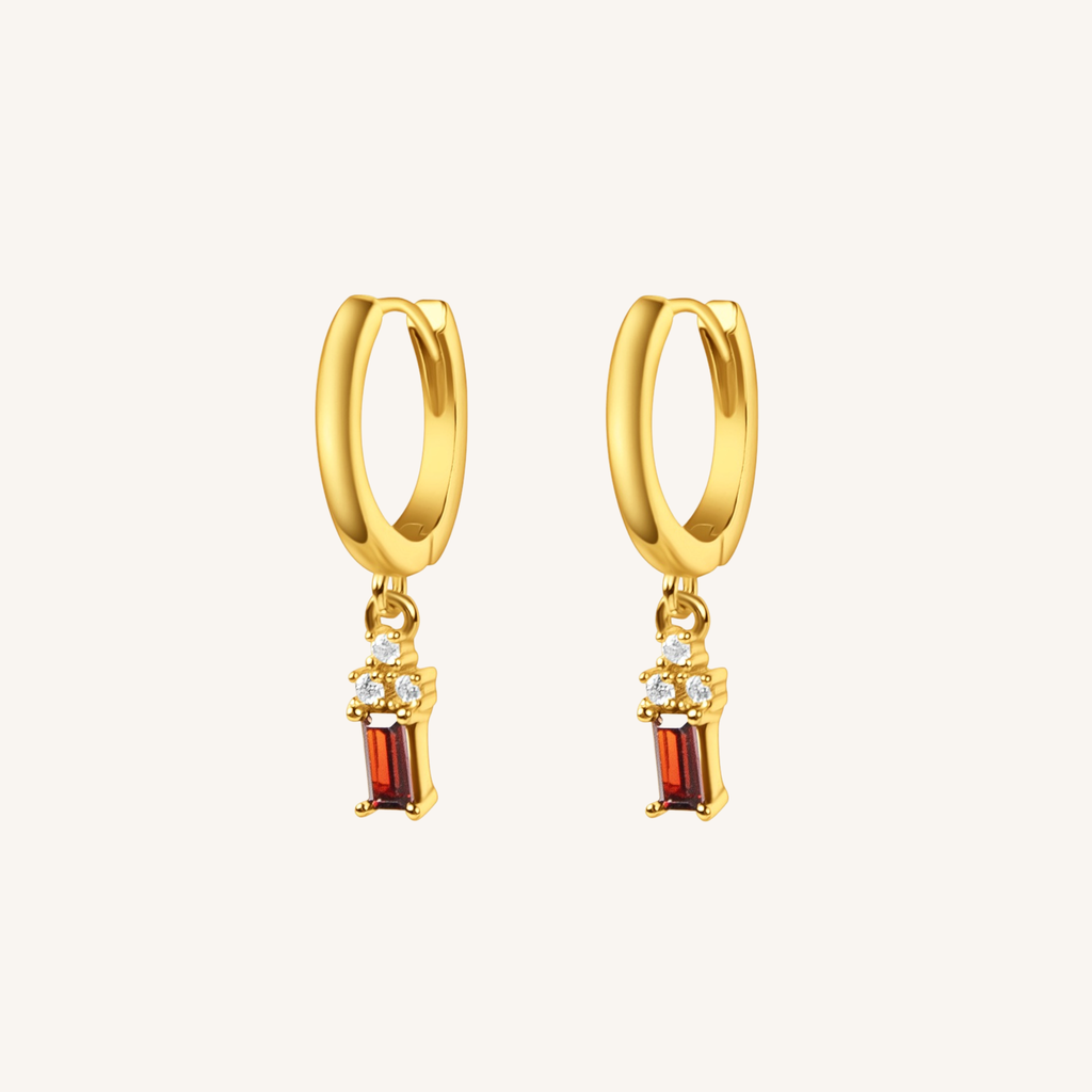 A pair of gold huggie hoop earrings with dangling rectangular red gemstone charms and small clear stones, shown on a light cream background.