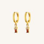 A pair of gold huggie hoop earrings with dangling rectangular red gemstone charms and small clear stones, shown on a light cream background.