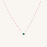A delicate rose gold chain necklace featuring a small square pendant with a vibrant green center, displayed against a solid off-white background.