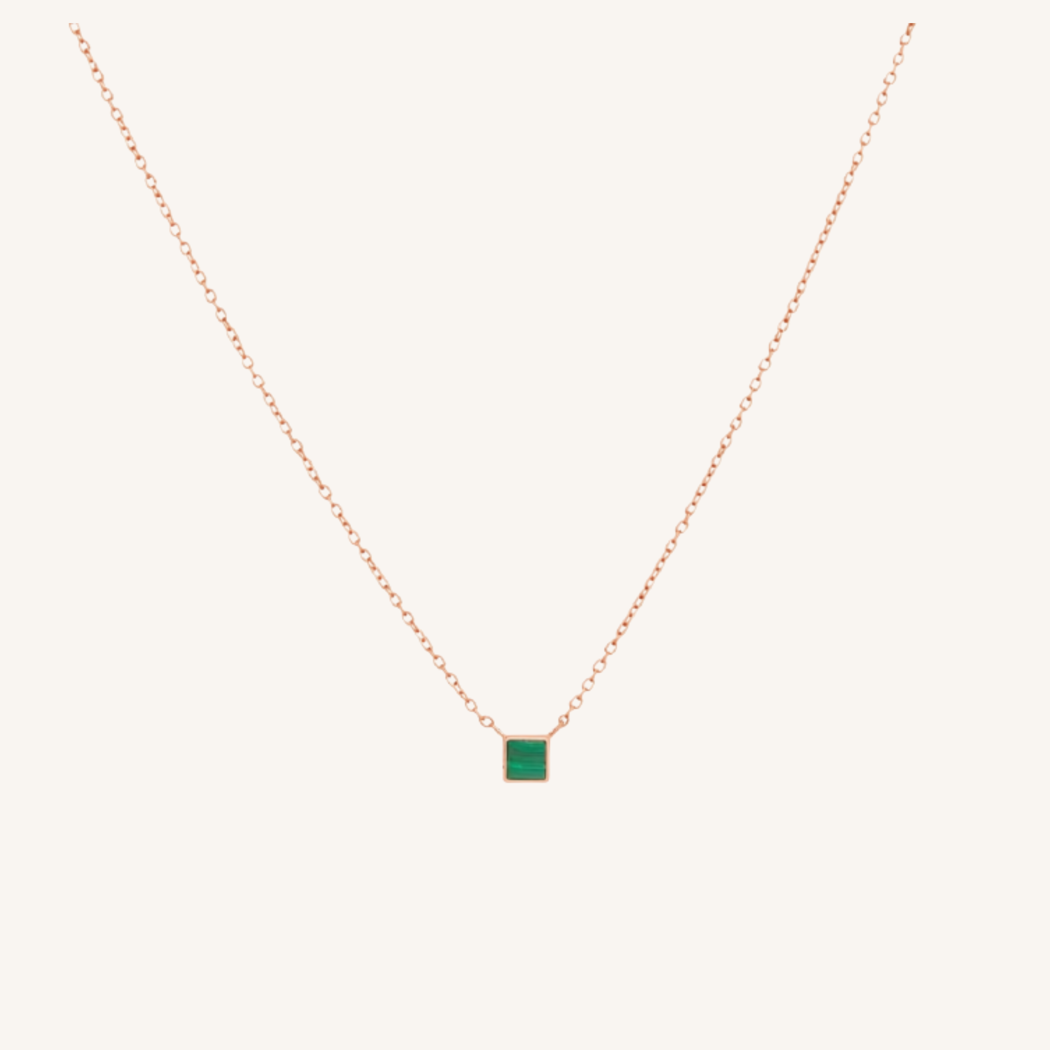 A delicate rose gold chain necklace featuring a small square pendant with a vibrant green center, displayed against a solid off-white background.