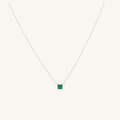 A delicate silver chain necklace featuring a small square green gemstone pendant, displayed against a solid off-white background.