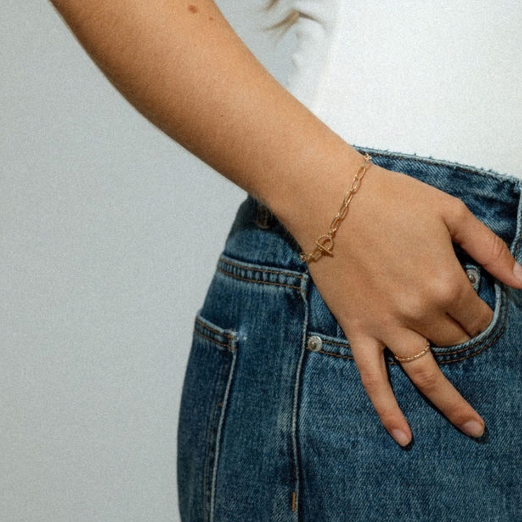 A close-up of a person's hand resting in the pocket of blue jeans, wearing a delicate gold chain bracelet with a small heart-shaped charm.