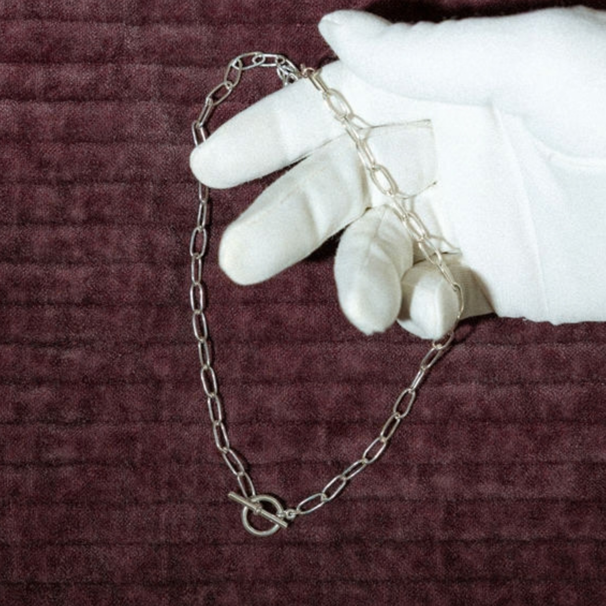 A silver-toned paperclip chain necklace with a toggle clasp, held by a white gloved hand against a textured dark burgundy background.