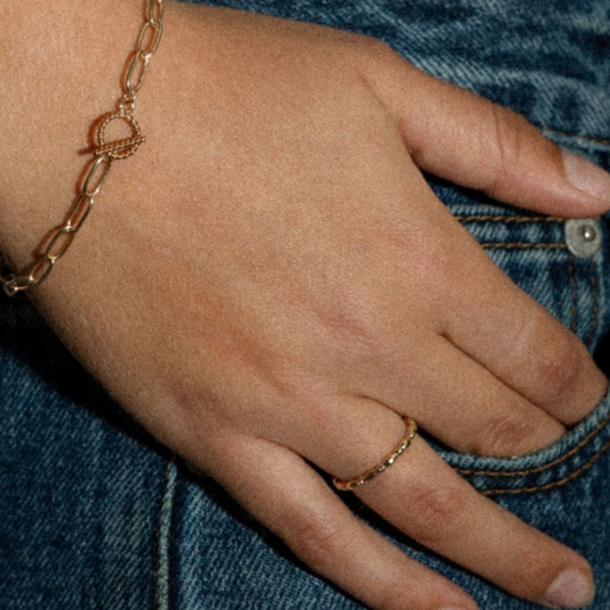 A close-up of a person's hand resting on blue denim jeans, wearing a delicate gold chain bracelet with a small heart-shaped link detail on the wrist and a matching thin gold ring on the finger.