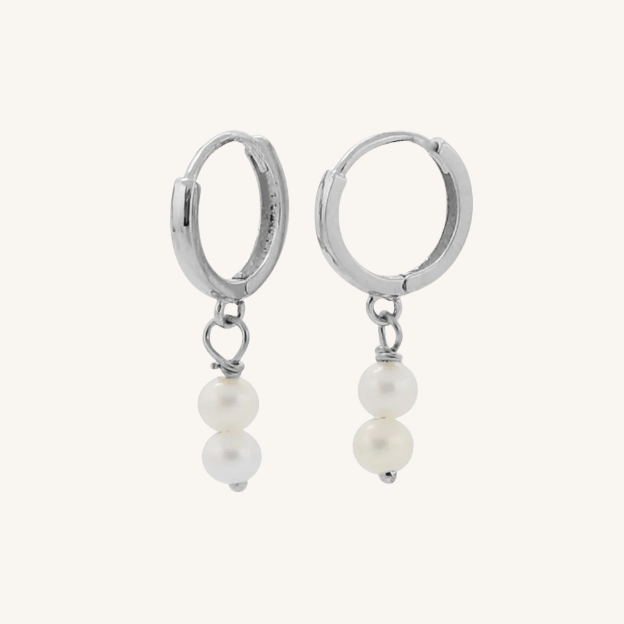 A pair of silver huggie hoop earrings with a dangling double pearl charm, shown against a solid off-white background.