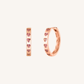 A pair of rose gold huggie hoop earrings decorated with small pink heart-shaped crystals, shown on a light cream background.