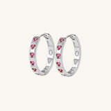 A pair of silver hoop earrings with small pink heart-shaped crystals set along the outer band, shown against a solid off-white background.