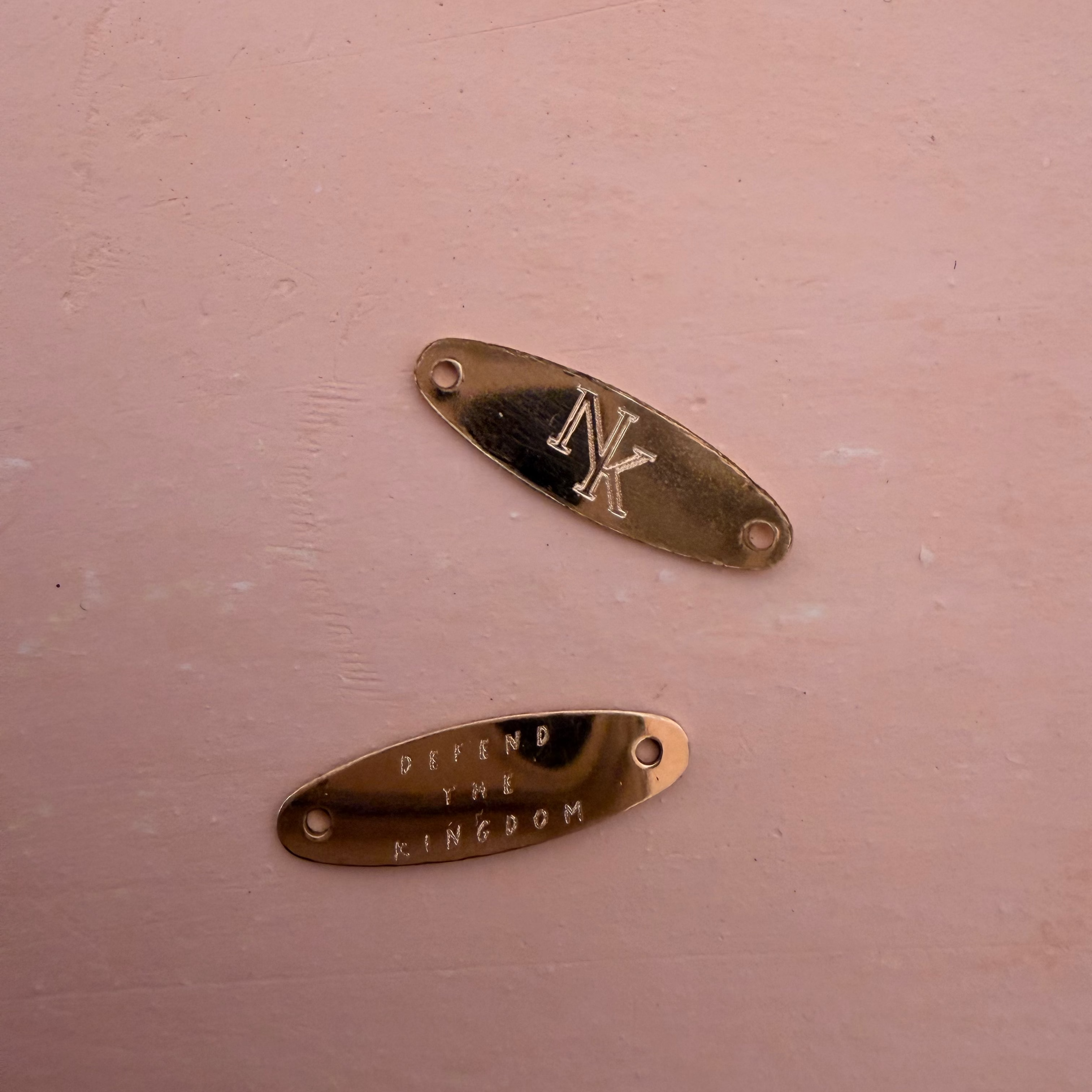 Two small metallic oval badges or plates on a dusty pink background. The top plate is gold-toned with an engraved stylized 'NK' monogram, and the bottom plate is bronze-toned with the engraved text 'OFFEND THE KINGDOM'.