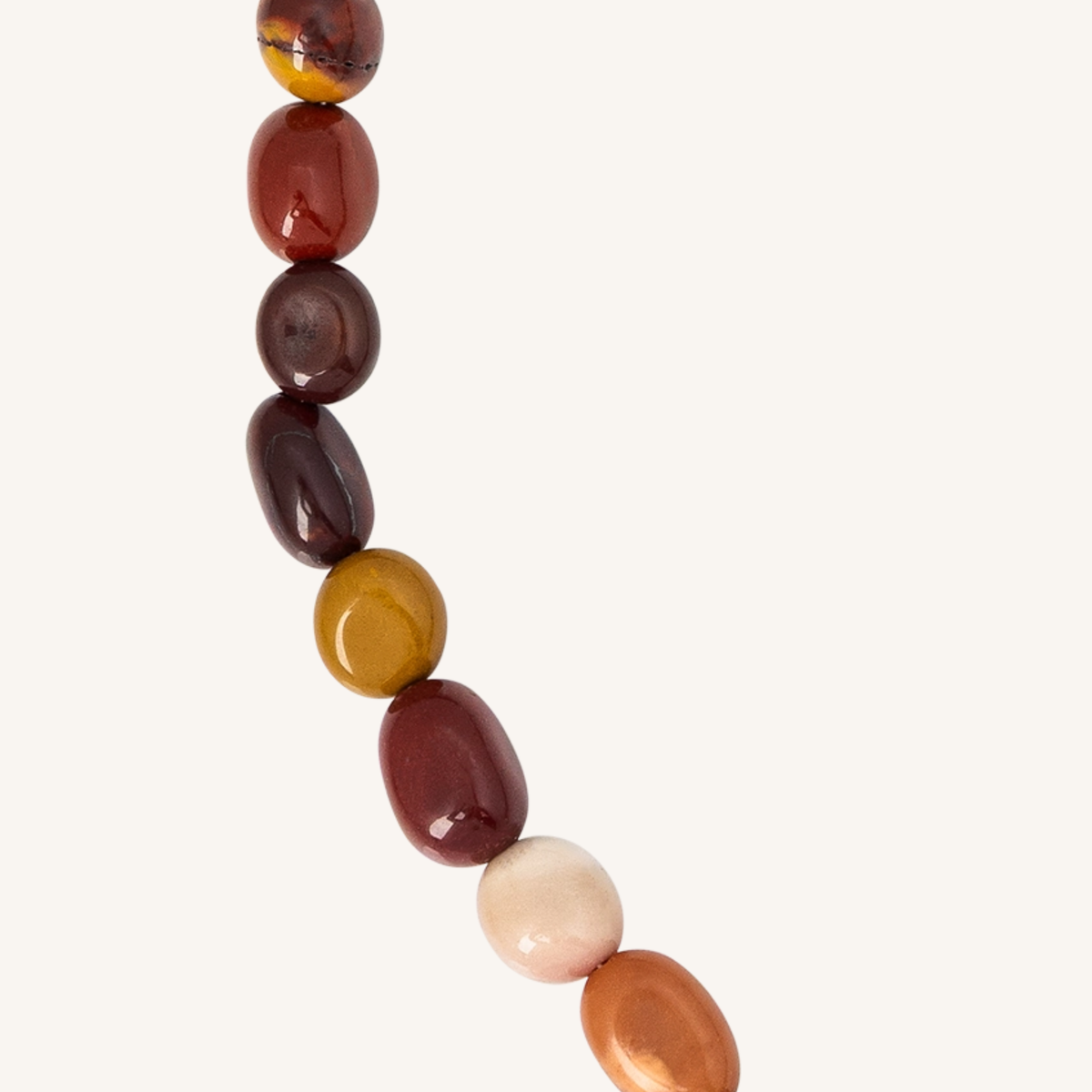 A close-up of a beaded strand featuring polished, smooth oval beads in various earthy colors including reddish-brown, dark brown, mustard yellow, peach, and cream, set against a solid off-white background.