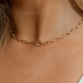 A close-up of a delicate gold chain necklace worn around a person's neck, featuring elongated oval links and a central textured circular ring clasp.
