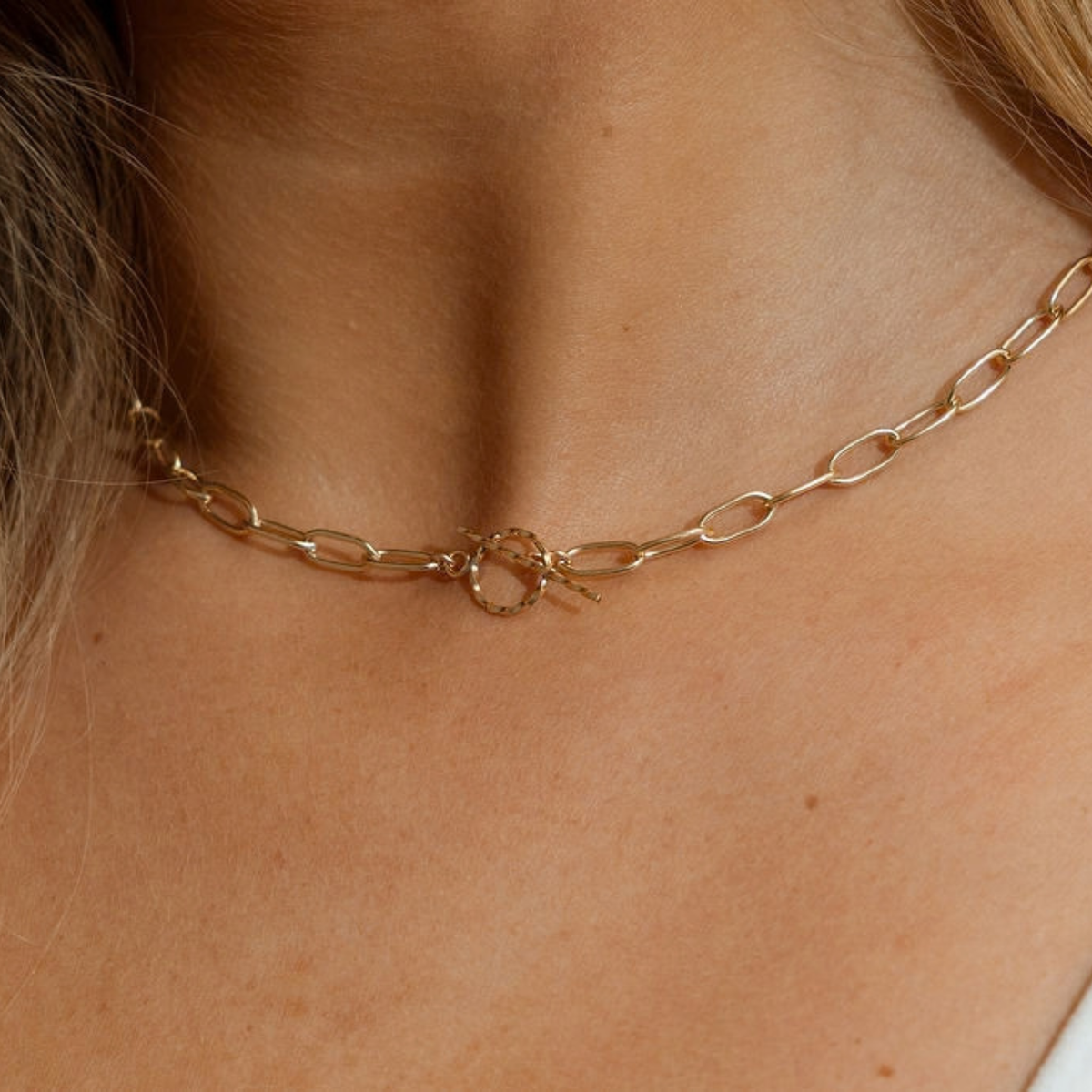 A close-up of a delicate gold chain necklace worn around a person's neck, featuring elongated oval links and a central textured circular ring clasp.