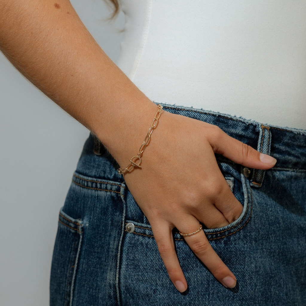A close-up of a person's hand resting in a blue denim pocket, wearing a delicate gold chain bracelet with a small heart-shaped pendant on the wrist.