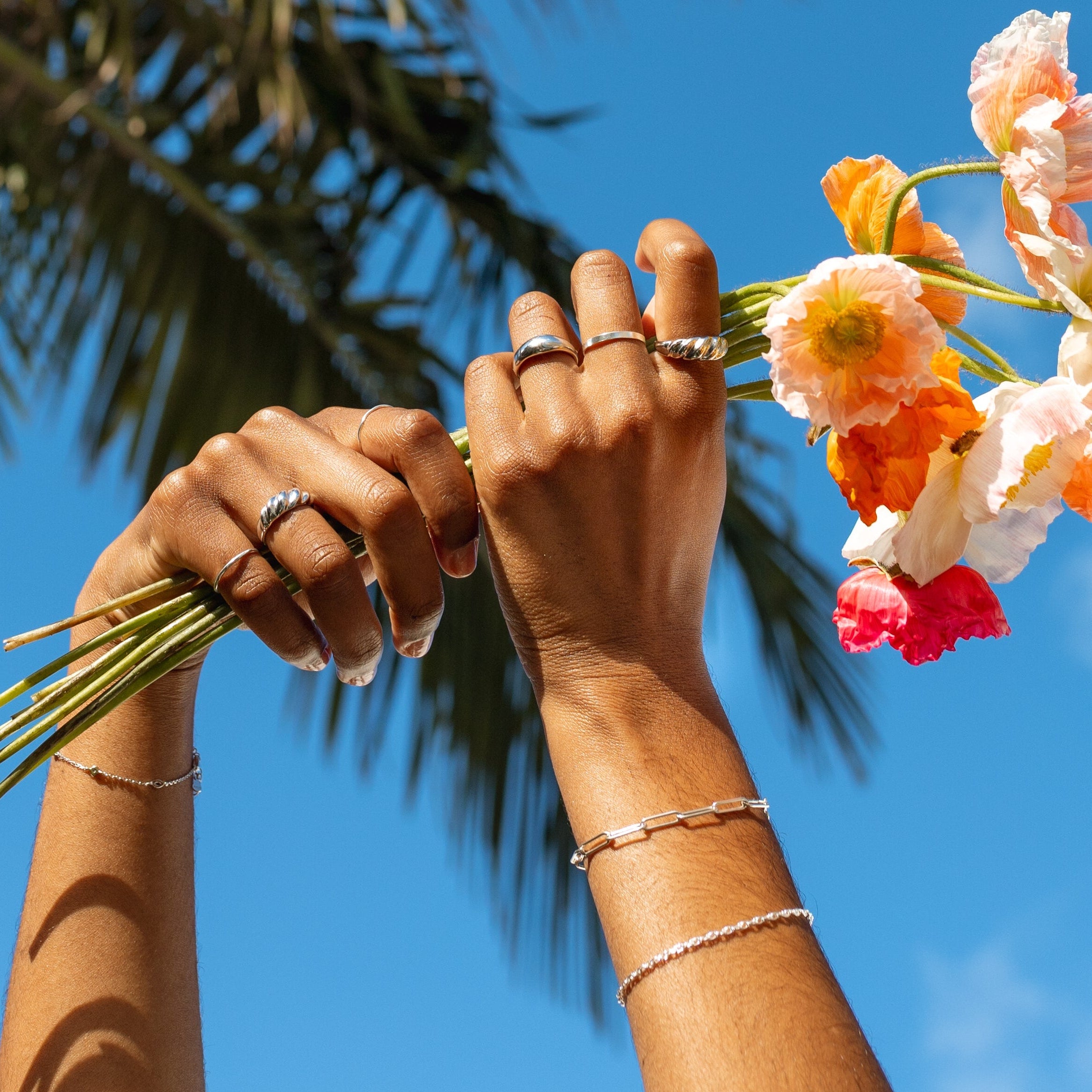 Two hands holding a bouquet of flowers against a palm tree and blue sky.