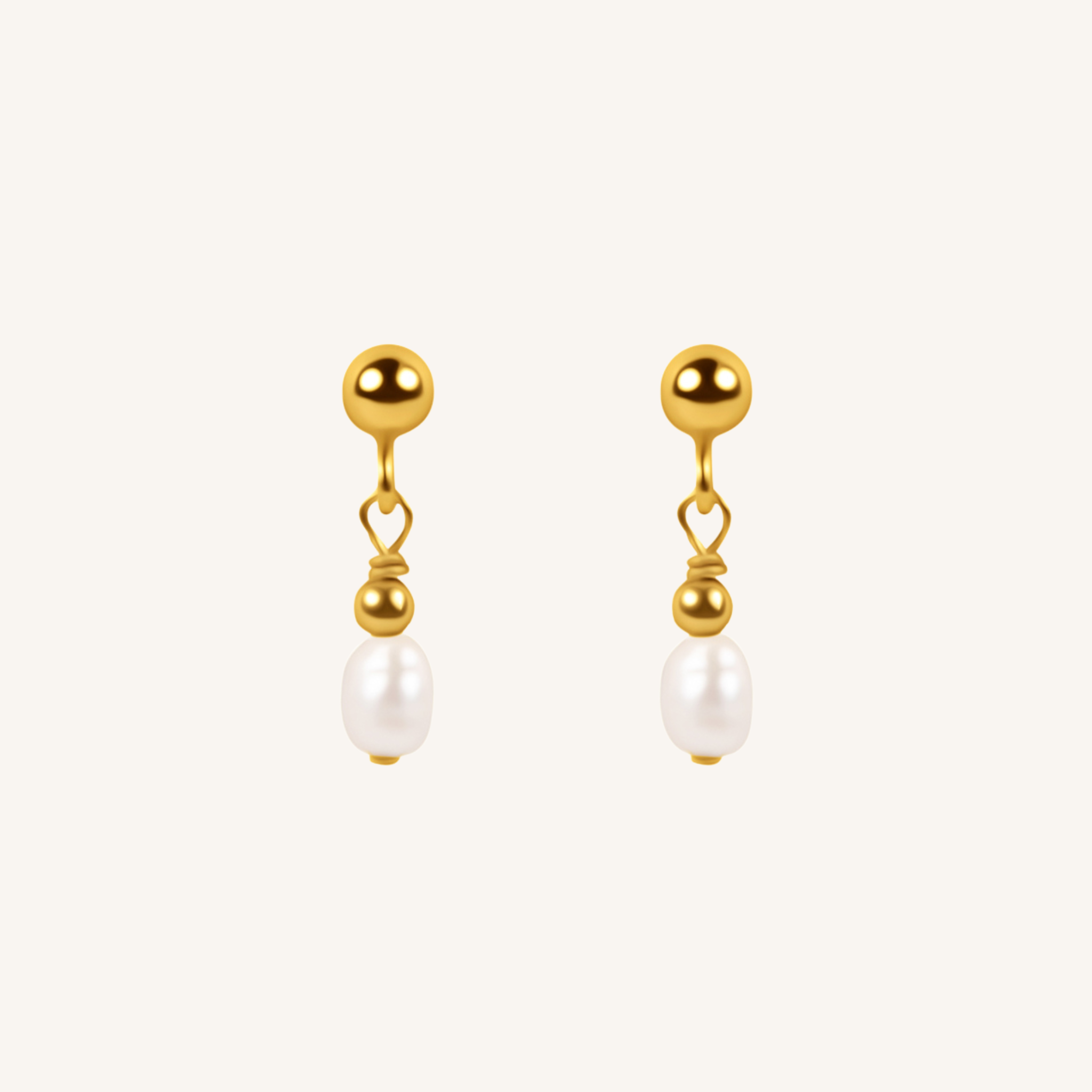 A pair of gold dangle earrings with a small heart-shaped wire detail and a white pearl drop, shown on a light cream background.