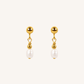 A pair of gold dangle earrings with a small heart-shaped wire detail and a white pearl drop, shown on a light cream background.