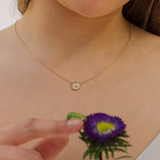 Gold necklace with a small round pendant worn by a person holding a purple flower.