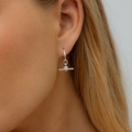 A close-up of a woman's ear wearing a delicate silver huggie hoop earring with a small horizontal bar and a tiny clear crystal charm.