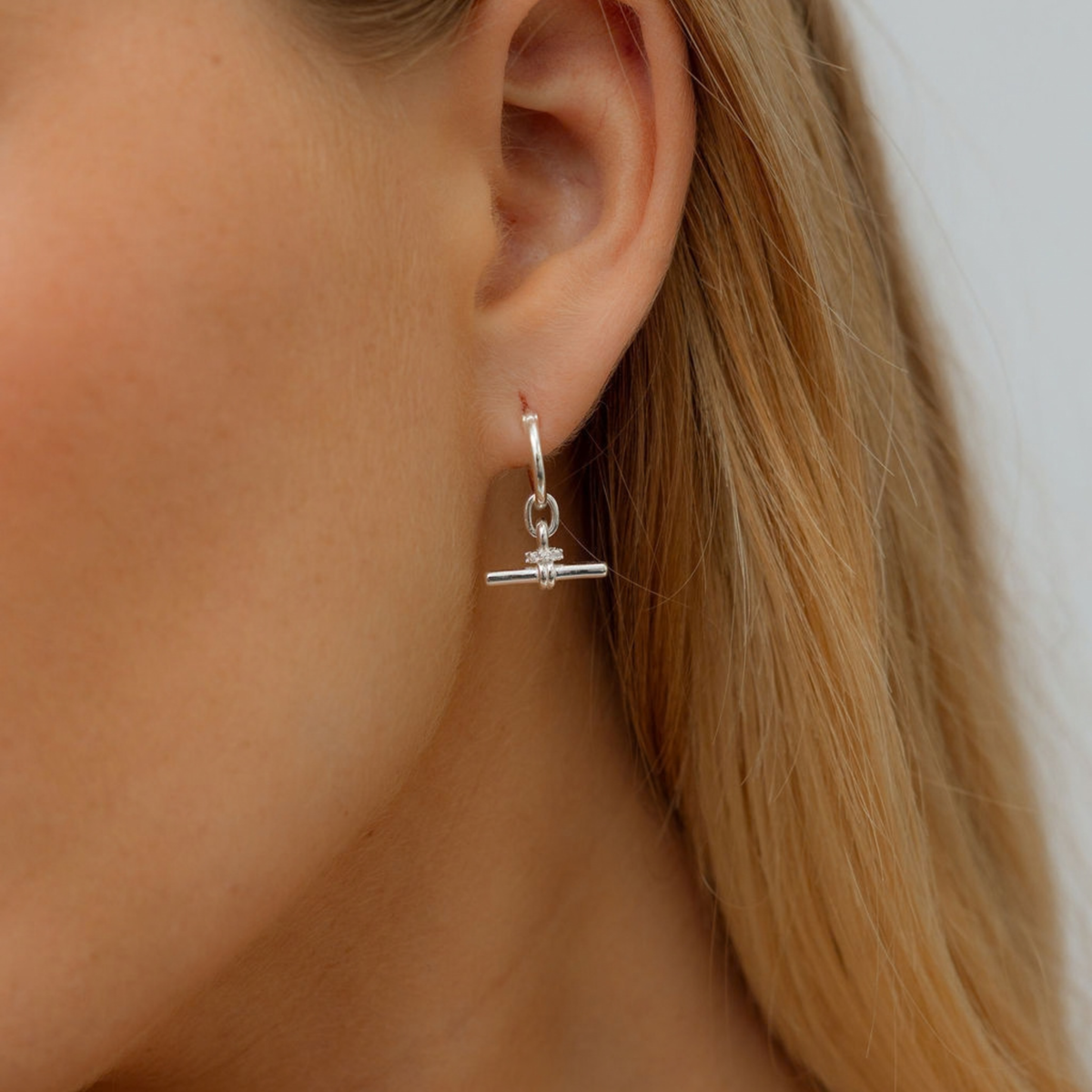 A close-up of a woman's ear wearing a delicate silver huggie hoop earring with a small horizontal bar and a tiny clear crystal charm.
