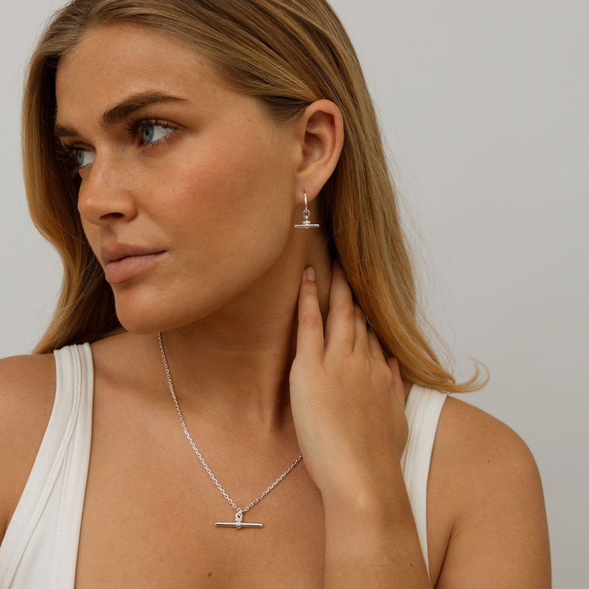 A woman wearing a delicate silver necklace with a horizontal bar pendant and matching drop earrings, shown in a close-up portrait against a light gray background.