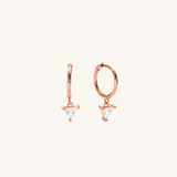 A pair of rose gold huggie hoop earrings with small triangular clear crystal drops, shown on a solid off-white background.