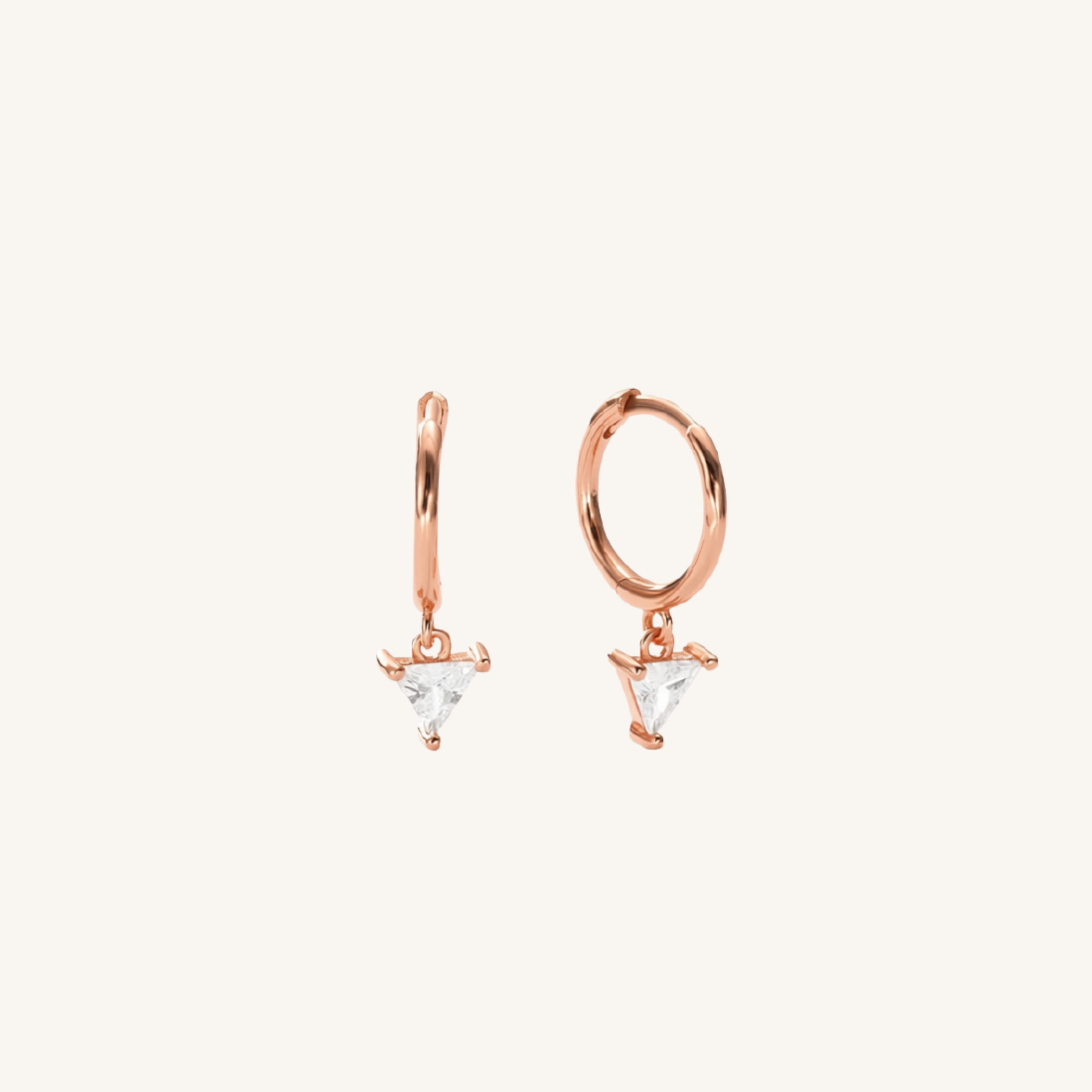 A pair of rose gold huggie hoop earrings with small triangular clear crystal drops, shown on a solid off-white background.