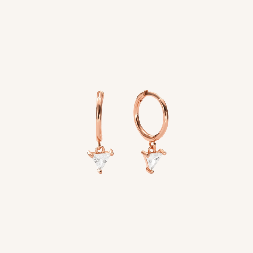 A pair of rose gold huggie hoop earrings with small triangular clear crystal drops, shown on a solid off-white background.