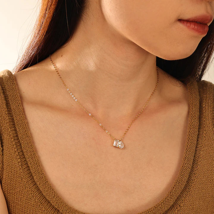 Model wearing a delicate gold chain necklace with a small pendant featuring two marquise-cut clear gemstones in a gold setting, displayed against a solid off-white background.