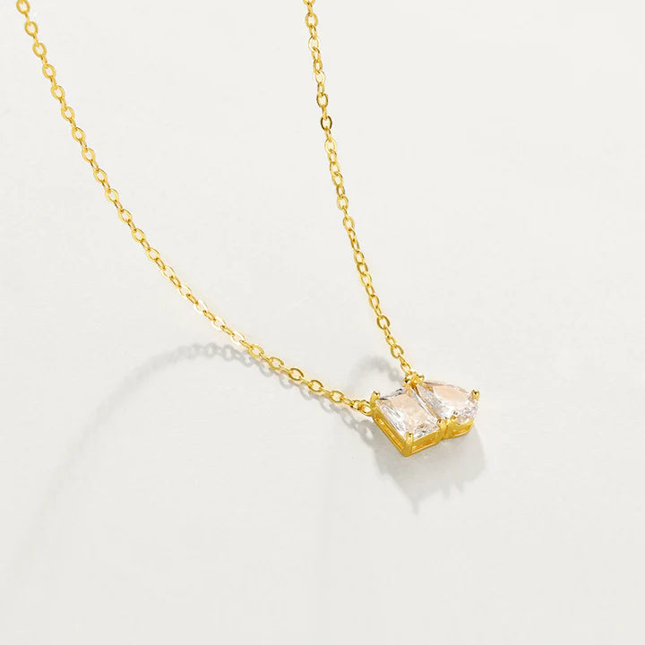 A delicate gold chain necklace with a small pendant featuring two marquise-cut clear gemstones in a gold setting, displayed against a solid off-white background.