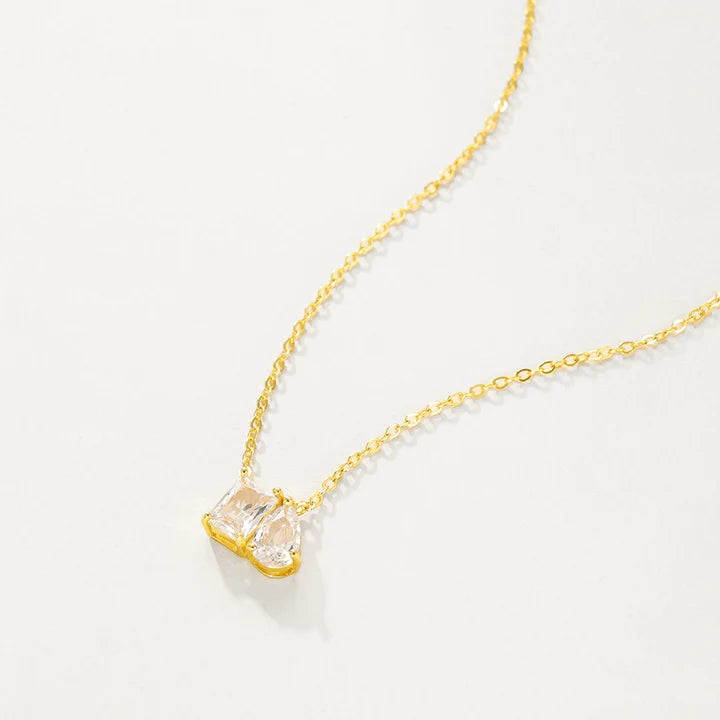 A delicate gold chain necklace with a small pendant featuring two marquise-cut clear gemstones in a gold setting, displayed against a solid off-white background.