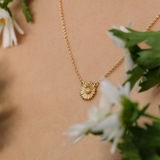 Gold necklace with a sunflower pendant worn by a person, surrounded by white flowers and green leaves.