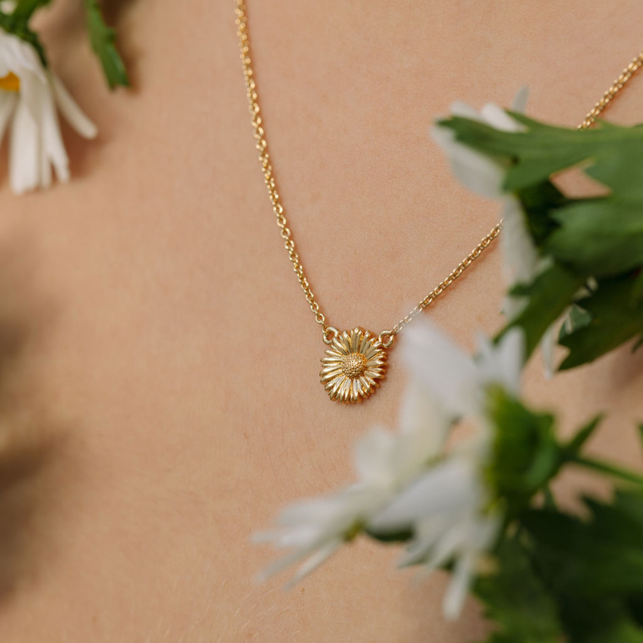 Gold necklace with a sunflower pendant worn by a person, surrounded by white flowers and green leaves.