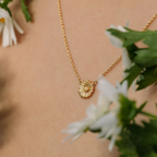 Gold necklace with a sunflower pendant worn by a person, surrounded by white flowers and green leaves.