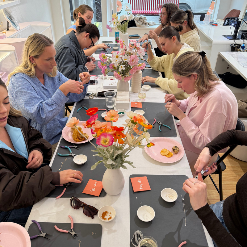 A group of women gathered around a table in a bright studio, making jewelry with pliers and small beads. The table is decorated with flower arrangements, plates with finished pieces, and orange cards.